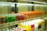 Rows of colorful beverage cans and bottles are neatly arranged on shelves inside a refrigerated display. The top shelf features green, red, and orange cans, while the bottom shelf holds yellow, pink, and white bottles.