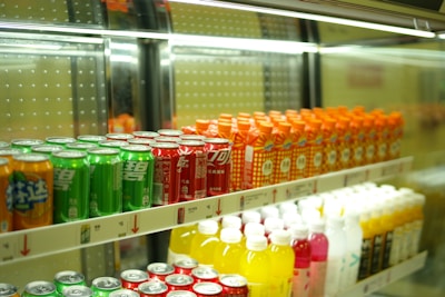 Rows of colorful beverage cans and bottles are neatly arranged on shelves inside a refrigerated display. The top shelf features green, red, and orange cans, while the bottom shelf holds yellow, pink, and white bottles.