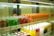 Rows of colorful beverage cans and bottles are neatly arranged on shelves inside a refrigerated display. The top shelf features green, red, and orange cans, while the bottom shelf holds yellow, pink, and white bottles.