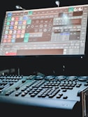 A dimly lit control desk with numerous buttons and knobs, paired with a large screen displaying complex software. The screen features a variety of icons and options related to lighting or stage effects.