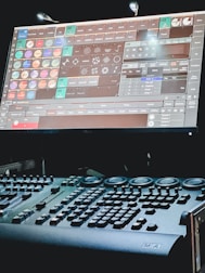 A dimly lit control desk with numerous buttons and knobs, paired with a large screen displaying complex software. The screen features a variety of icons and options related to lighting or stage effects.