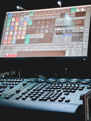 A dimly lit control desk with numerous buttons and knobs, paired with a large screen displaying complex software. The screen features a variety of icons and options related to lighting or stage effects.