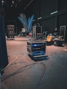 A backstage area containing various technical equipment such as audio racks, flight cases, and cables spread across the floor. The dimly lit environment suggests preparation for an event. Brightly colored banners with text and a few scattered chairs can be seen in the background.