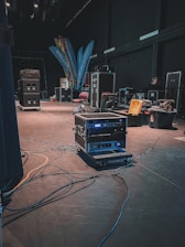 A backstage area containing various technical equipment such as audio racks, flight cases, and cables spread across the floor. The dimly lit environment suggests preparation for an event. Brightly colored banners with text and a few scattered chairs can be seen in the background.