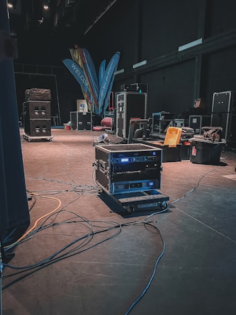 A backstage area containing various technical equipment such as audio racks, flight cases, and cables spread across the floor. The dimly lit environment suggests preparation for an event. Brightly colored banners with text and a few scattered chairs can be seen in the background.
