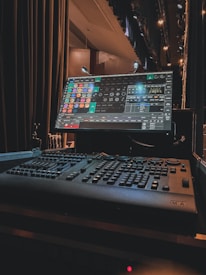 A lighting control console is prominently featured with a digital interface displaying various buttons and dials. The screen shows a colorful, detailed control panel for stage lighting. The surrounding environment includes stage curtains and lights, suggesting a theater or performance setting.
