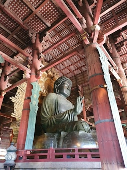 A large bronze statue of Buddha sits inside a historic wooden temple. The intricate wooden ceiling and architecture surrounding the statue include carved details and supporting pillars painted in red. The serene expression on the Buddha's face adds a spiritual ambiance to the setting.