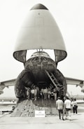 A large cargo aircraft with its nose lifted, revealing the vast interior cargo hold. Several people are walking up the ramp into the aircraft, while a 'No Smoking' sign is visible at the entrance. The scene takes place on an airfield with other aircraft parts visible in the background.