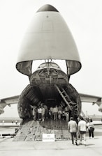 A large cargo aircraft with its nose lifted, revealing the vast interior cargo hold. Several people are walking up the ramp into the aircraft, while a 'No Smoking' sign is visible at the entrance. The scene takes place on an airfield with other aircraft parts visible in the background.