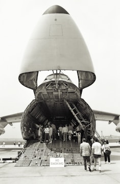 A large cargo aircraft with its nose lifted, revealing the vast interior cargo hold. Several people are walking up the ramp into the aircraft, while a 'No Smoking' sign is visible at the entrance. The scene takes place on an airfield with other aircraft parts visible in the background.