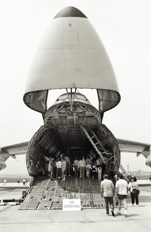 A large cargo aircraft with its nose lifted, revealing the vast interior cargo hold. Several people are walking up the ramp into the aircraft, while a 'No Smoking' sign is visible at the entrance. The scene takes place on an airfield with other aircraft parts visible in the background.