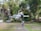 A white drone hovers in the air, captured sharply against a blurred background of trees and buildings. The drone's design features four propellers, and it has a compact, modern appearance with the model name 'MINI 3 PRO' visible on its side.