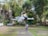 A white drone hovers in the air, captured sharply against a blurred background of trees and buildings. The drone's design features four propellers, and it has a compact, modern appearance with the model name 'MINI 3 PRO' visible on its side.