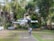 A white drone hovers in the air, captured sharply against a blurred background of trees and buildings. The drone's design features four propellers, and it has a compact, modern appearance with the model name 'MINI 3 PRO' visible on its side.