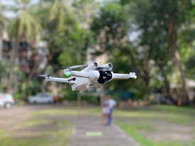 A white drone hovers in the air, captured sharply against a blurred background of trees and buildings. The drone's design features four propellers, and it has a compact, modern appearance with the model name 'MINI 3 PRO' visible on its side.