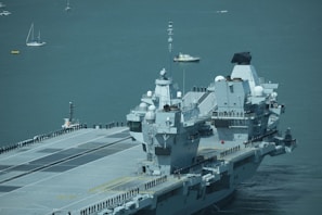 A large aircraft carrier is sailing on a calm sea with uniformed personnel lined up along the deck. Several smaller boats and sailboats are in the background.