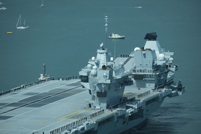 A large aircraft carrier is sailing on a calm sea with uniformed personnel lined up along the deck. Several smaller boats and sailboats are in the background.