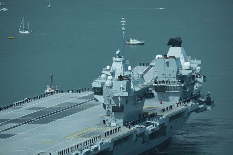 A large aircraft carrier is sailing on a calm sea with uniformed personnel lined up along the deck. Several smaller boats and sailboats are in the background.