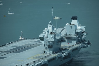A large aircraft carrier is sailing on a calm sea with uniformed personnel lined up along the deck. Several smaller boats and sailboats are in the background.