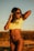 A woman is posing outdoors during sunset, wearing sunglasses, a yellow Brazil soccer jersey, and black swimwear bottoms. Her hair is flowing in the wind.