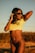 A woman is posing outdoors during sunset, wearing sunglasses, a yellow Brazil soccer jersey, and black swimwear bottoms. Her hair is flowing in the wind.