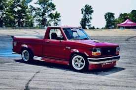 A modified red pickup truck is performing a drift on a racetrack. The truck is equipped with custom rims, a spoiler, and various decals on the body. Smoke is visible near the rear wheels, indicating the drifting action. Trees and a tent are in the background, suggesting an outdoor setting.