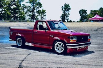 A modified red pickup truck is performing a drift on a racetrack. The truck is equipped with custom rims, a spoiler, and various decals on the body. Smoke is visible near the rear wheels, indicating the drifting action. Trees and a tent are in the background, suggesting an outdoor setting.