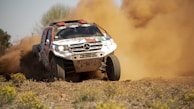 A rally car speeds across a dusty off-road track, kicking up a large cloud of dirt and dust behind it. Wildflowers and sparse grass are visible in the foreground, adding lively color to the otherwise dry landscape. The car is aggressively styled with decals, emphasizing its competitive nature.
