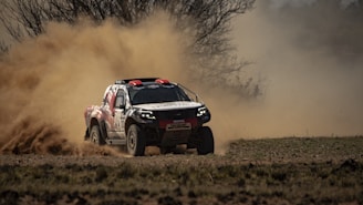 A rally car speeds through a dirt path, kicking up a cloud of dust behind it. The background shows barren trees and a hazy sky, indicating a rugged and outdoor setting.