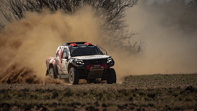 A rally car speeds through a dirt path, kicking up a cloud of dust behind it. The background shows barren trees and a hazy sky, indicating a rugged and outdoor setting.