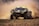A high-performance off-road race car speeds over a dusty terrain, kicking up clouds of dirt. The car is covered in various sponsor logos, has a sleek design with distinctive green and black coloring, and large rugged tires suitable for rough surfaces.