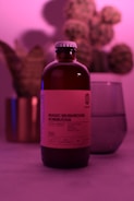 A bottle labeled 'Magic Mushroom Kombucha' is prominently featured, surrounded by a soft, pinkish-purple ambient light. In the background, there is a blurred image of a plant and a glass object, possibly a cup or a vase. The overall scene is moody and uses diffused lighting to create a calming atmosphere.