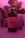 A bottle labeled 'Magic Mushroom Kombucha' is prominently featured, surrounded by a soft, pinkish-purple ambient light. In the background, there is a blurred image of a plant and a glass object, possibly a cup or a vase. The overall scene is moody and uses diffused lighting to create a calming atmosphere.