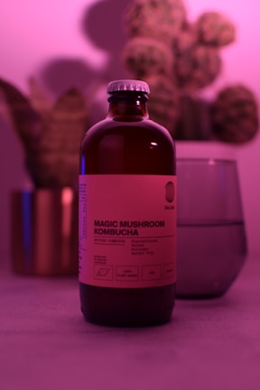 A bottle labeled 'Magic Mushroom Kombucha' is prominently featured, surrounded by a soft, pinkish-purple ambient light. In the background, there is a blurred image of a plant and a glass object, possibly a cup or a vase. The overall scene is moody and uses diffused lighting to create a calming atmosphere.