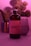 A bottle labeled 'Magic Mushroom Kombucha' is prominently featured, surrounded by a soft, pinkish-purple ambient light. In the background, there is a blurred image of a plant and a glass object, possibly a cup or a vase. The overall scene is moody and uses diffused lighting to create a calming atmosphere.