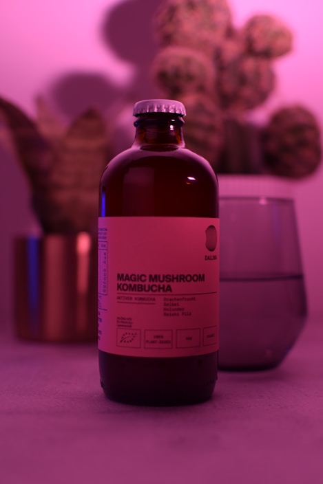 A bottle labeled 'Magic Mushroom Kombucha' is prominently featured, surrounded by a soft, pinkish-purple ambient light. In the background, there is a blurred image of a plant and a glass object, possibly a cup or a vase. The overall scene is moody and uses diffused lighting to create a calming atmosphere.