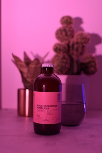A bottle of Magic Mushroom Kombucha is centered on a surface, illuminated by pink and purple lighting. In the background, there is a glass partially filled with a beverage and two plants, including a cactus, creating a relaxed and modern ambiance.