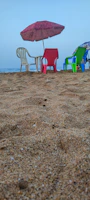 Several colorful plastic chairs arranged on a sandy beach with a red umbrella providing shade. The ocean is visible in the background, creating a tranquil seaside setting.