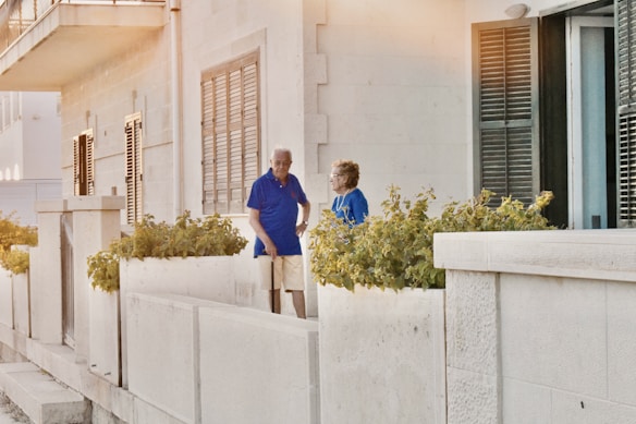An elderly man and woman stand on a terrace next to a building with beige stone walls and shuttered windows. They are surrounded by potted plants with green foliage. The man is wearing a blue shirt and khaki shorts, while the woman is dressed in a blue blouse. The atmosphere has a warm, late-afternoon lighting.