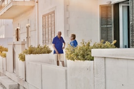 An elderly man and woman stand on a terrace next to a building with beige stone walls and shuttered windows. They are surrounded by potted plants with green foliage. The man is wearing a blue shirt and khaki shorts, while the woman is dressed in a blue blouse. The atmosphere has a warm, late-afternoon lighting.