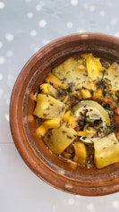 A tagine dish featuring various roasted vegetables, including yellow squash, carrots, and a partially squeezed lime, topped with herbs. The food is placed in a traditional earthenware pot, and the image is lit with dappled sunlight creating a playful pattern.
