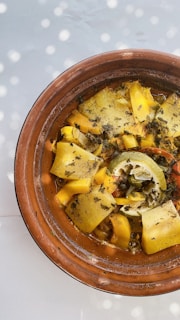 A tagine dish featuring various roasted vegetables, including yellow squash, carrots, and a partially squeezed lime, topped with herbs. The food is placed in a traditional earthenware pot, and the image is lit with dappled sunlight creating a playful pattern.
