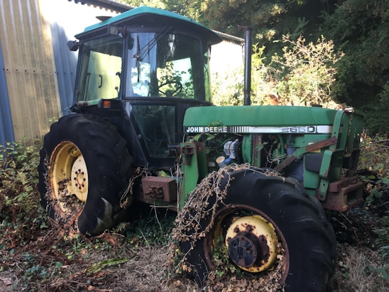 An old, green John Deere tractor is parked outdoors near a sheet metal building. The tractor appears weathered, with vegetation growing around its wheels, suggesting it has been stationary for some time. The setting includes overgrown bushes and trees, indicating it is in a rural or farm environment.