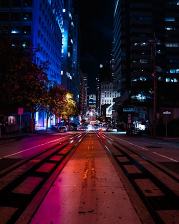 A vibrant urban street at night with bright neon lights illuminating the surroundings. Tall buildings on either side create a corridor effect, while the street lights cast colorful reflections on the pavement. The scene features a perspective view of a long street with visible tram tracks.