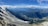 A breathtaking aerial view of a vast mountain range with a prominent glacier winding through the landscape. The foreground features rugged, snow-capped peaks, while a dense valley stretches out below. The sky is bright blue, dotted with fluffy white clouds.