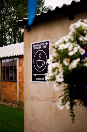 A cozy outdoor setting features a sign on a wall indicating the entrance to Winstan's Bunker Cafe, adorned with a steaming coffee cup graphic. In the foreground, a vibrant hanging flower basket with white blossoms adds a touch of charm. The background reveals part of a rustic building with large windows, framed by greenery and trees.