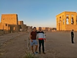 Two people stand in front of ancient Egyptian ruins featuring tall columns and hieroglyphs. The sky is clear with a warm tone during what appears to be either sunrise or sunset, casting a golden glow over the stone structures.