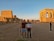 Two people stand in front of ancient Egyptian ruins featuring tall columns and hieroglyphs. The sky is clear with a warm tone during what appears to be either sunrise or sunset, casting a golden glow over the stone structures.