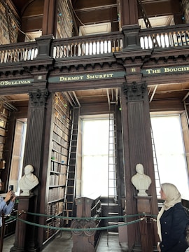 A grand library setting with tall, wooden bookshelves filled with numerous books. Busts of notable figures are displayed on either side of the entrance, adorned with green plaques bearing names. Two individuals are visible; one taking a photo and the other observing the surroundings. Ladders are positioned to provide access to higher shelves.