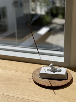 A serene setting featuring a light wooden surface by a window, with sunlight streaming in. An incense stick is burning, emitting a thin wisp of smoke. It is held upright on a stone base, which is adorned with a small sculpture of a reclining dog. The background through the window is blurred, giving a peaceful and contemplative feel to the scene.